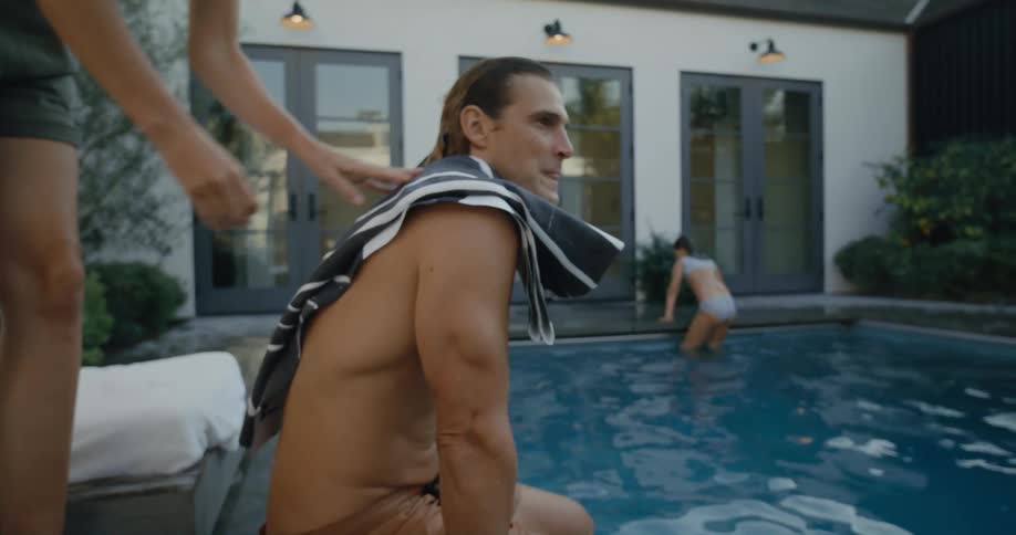 Shirtless athletic man sitting at the pool edge when a woman throws a towel on his back and sits behind him at the pool lounger