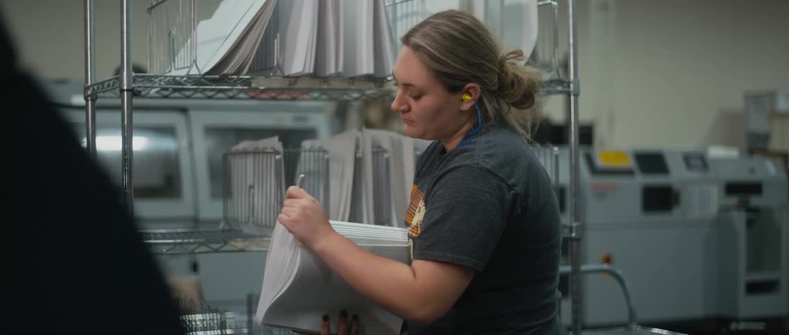 A woman working in a mail room writes on a stack of papers and places them in a cart