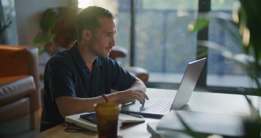 A man is sitting at a table using a laptop computer as the sun shines brightly in the window