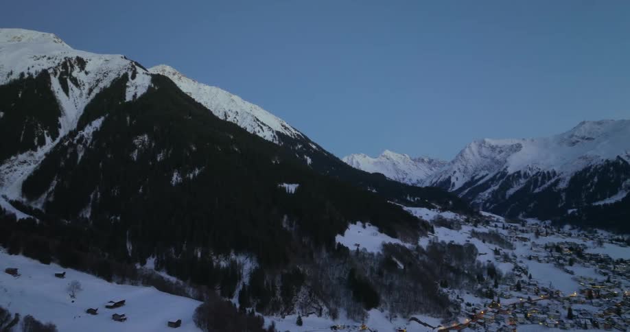 Aerial view of distant snowy town surrounded by snowy mountains at dusk
