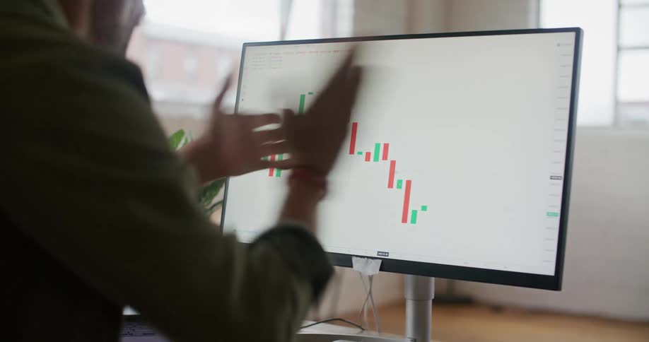 A man is sitting at a desk looking at a laptop computer with a graph on the screen and cheering