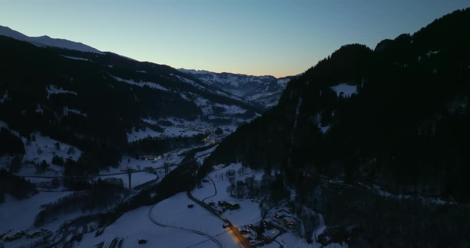 Automobiles travel on a forested mountain valley road surrounded by snow