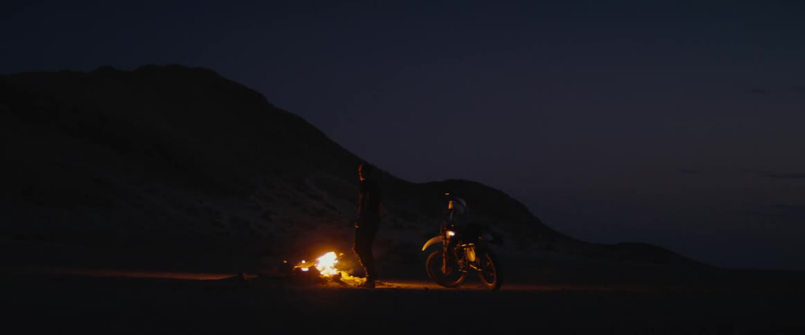 Man standing next to a campfire during the night and taking off his jacket to put it on his motorcycle parked next to him