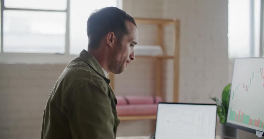 A man yawns while viewing stock charts on a computer at a desk indoors during the day