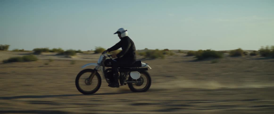 A man wearing a white helmet daringly drives his motorcycle across a dusty desert plain