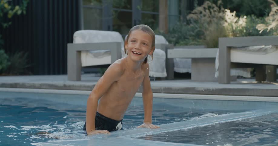 Smiling boy climbs out of swimming pool with outdoor furniture sitting between house and pool