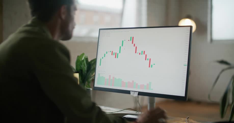 A man is sitting at a desk looking at a monitor with a graph on it and a laptop computer