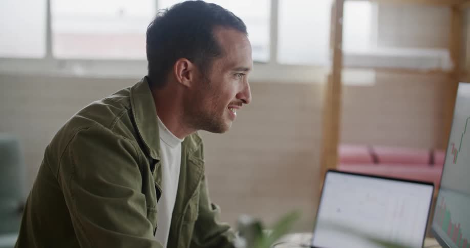 Man sitting at the table working on his laptop and monitor screen by analyzing a candle chart