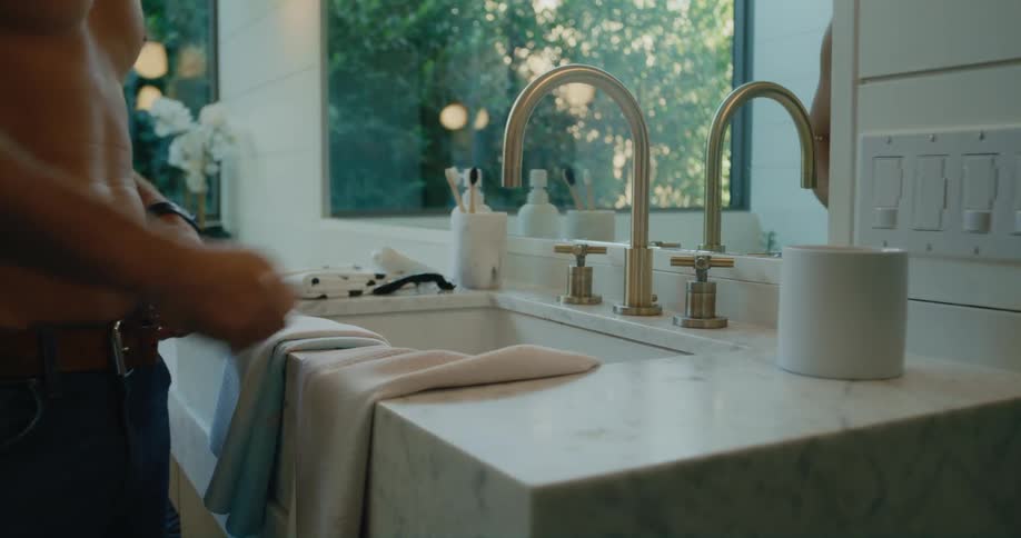 Shirtless man turning the bathroom tap off and leaving the towel at the sink