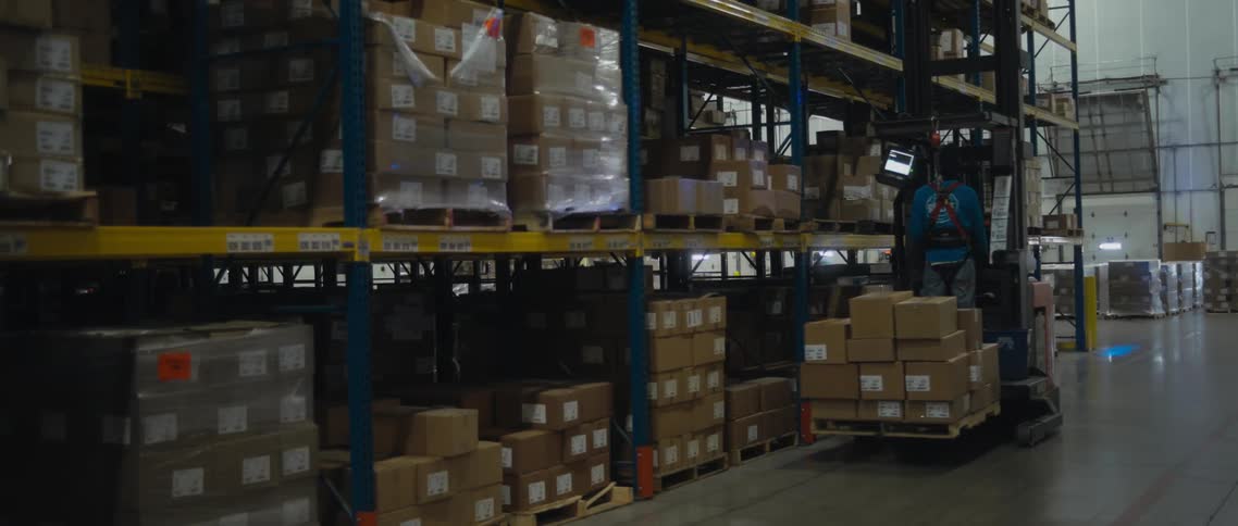 A man drives a lifter with boxes in a warehouse as another man pushes a cart filled with boxes