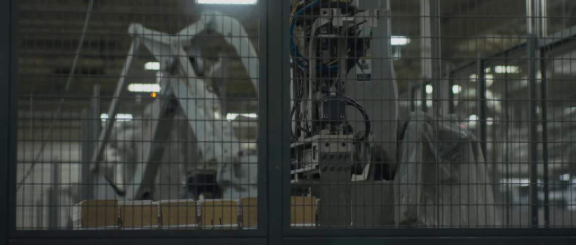Pair of robotic arms place down objects behind mesh enclosure in factory