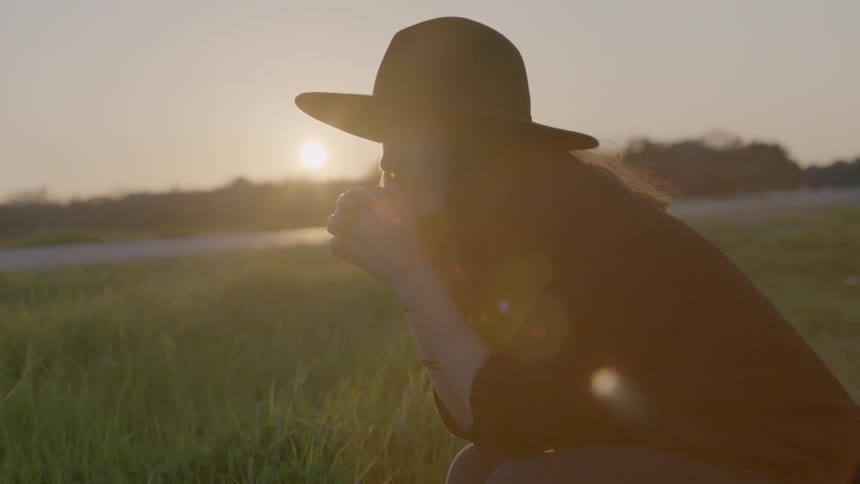 Woman in hat kneeling in a grassy field at sunset