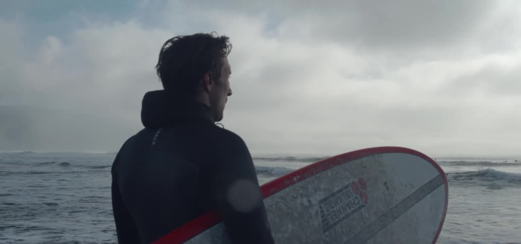 A man is standing near the water holding his surfboard and looking at the waves as a storm moves in