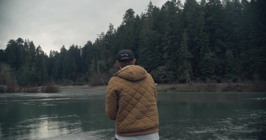 A man wearing a parka is standing next to a forest lake holding a fishing pole