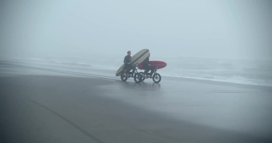 Two men riding motorcycles while holding surfboards in an empty beach during a fog day