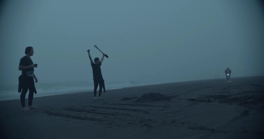 Friends cheer one holding a shovel as motorcyclist rides over sand mound on beach on a foggy day