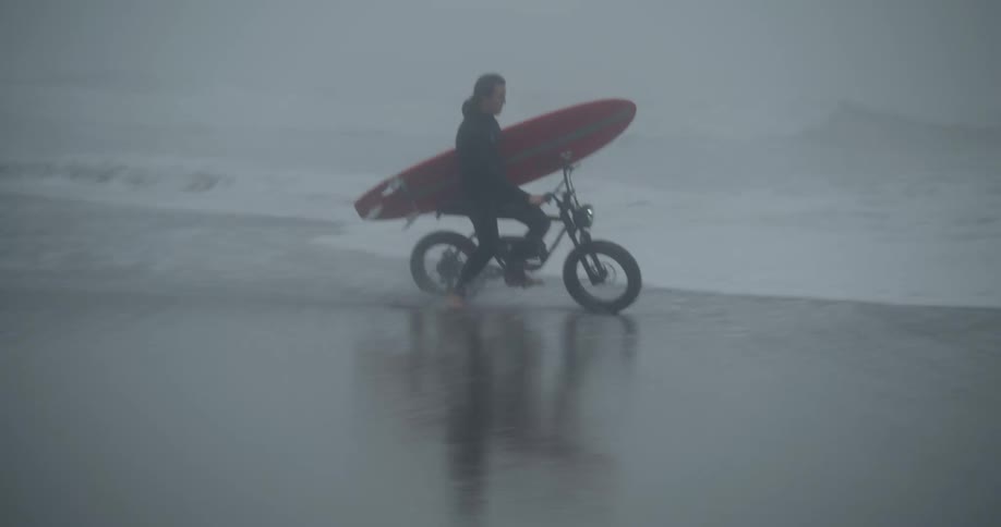 Man holding a surf board while riding a motorized bike next to the ocean in the beach at a rainy day