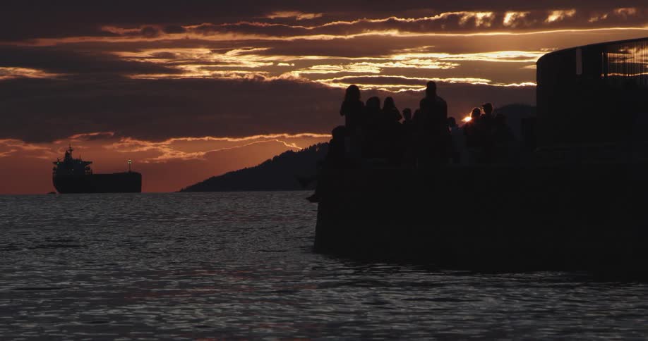 A group of people sitting on a seawall silhouetted against a red sky as freighter sits on the horizon