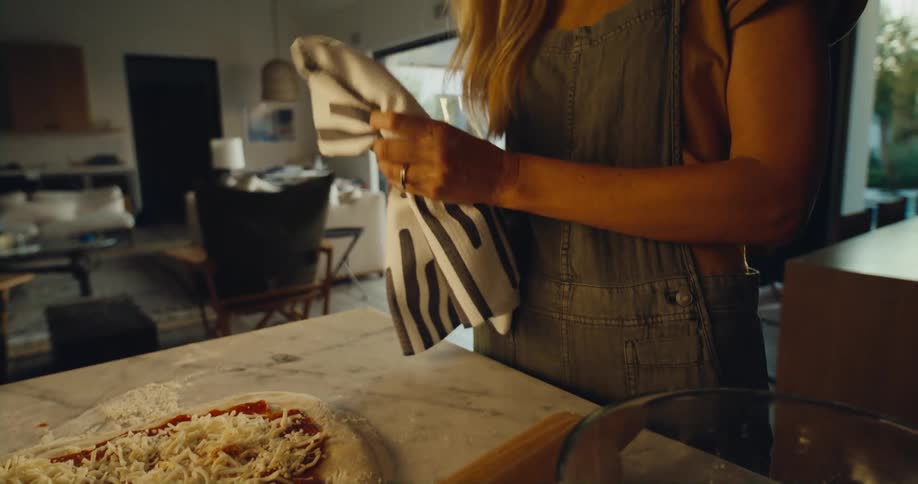 Woman standing in front of a kitchen counter inside a house preparing a pizza and cleaning her hand in a hand cloth