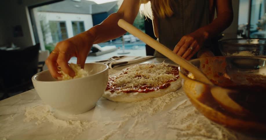 Woman preparing a pizza throwing cheese into the pizza dough at the kitchen counter inside the house
