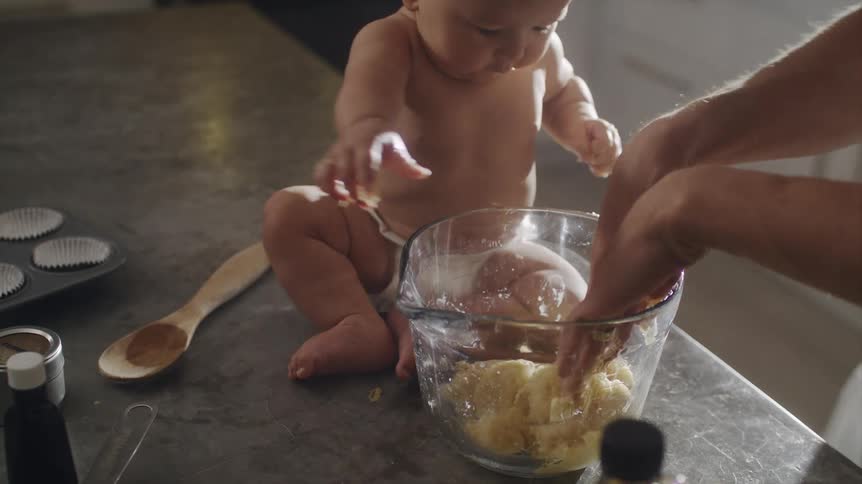 Baby wearing a chef hat helping dad mix dough in a glass bowl