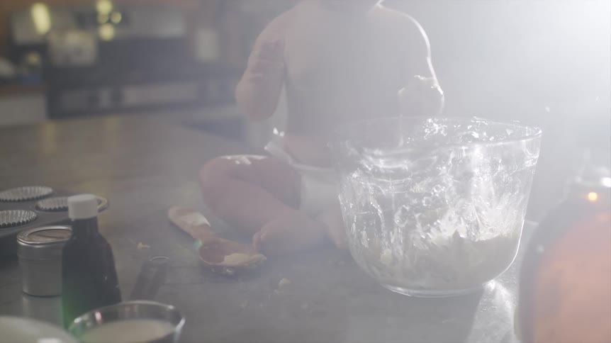 Child sits on kitchen counter eating as parent mixes mixture as sun glares