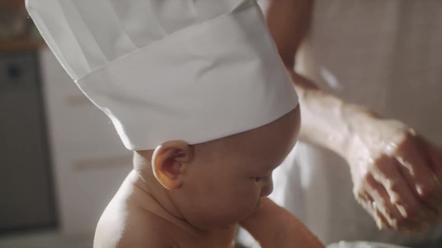 A baby sits on the kitchen counter and uses hands to help its father mix batter