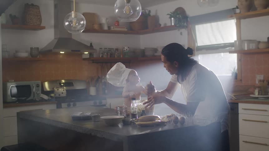 A man prepares food in a kitchen while a baby in a chef hat sits on the counter and watches