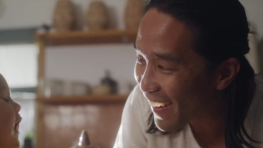 Father cooking in the kitchen while talking and laughingn with his baby son that is sitting next to him