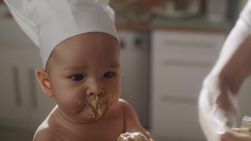 Baby wearing a chefs hat with his face and hands dirty with food sitting next to a bowl in which a man is mixing a dough