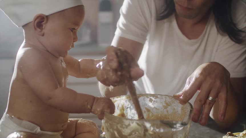 Asian man mixing food in a glass bowl with his baby wearing a chef hat sitting at the kitchen counter next to it