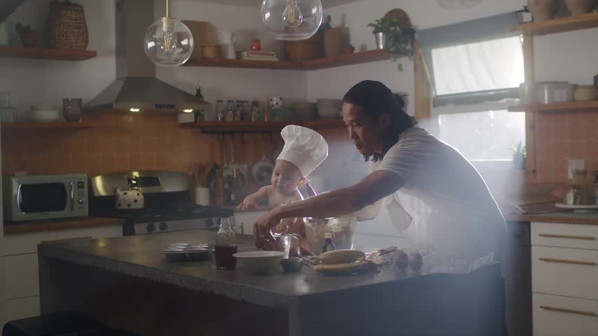 Father trying to cook on the kitchen counter and having to stop his baby son wearing a chef hat from making a mess with the kitchen utensils