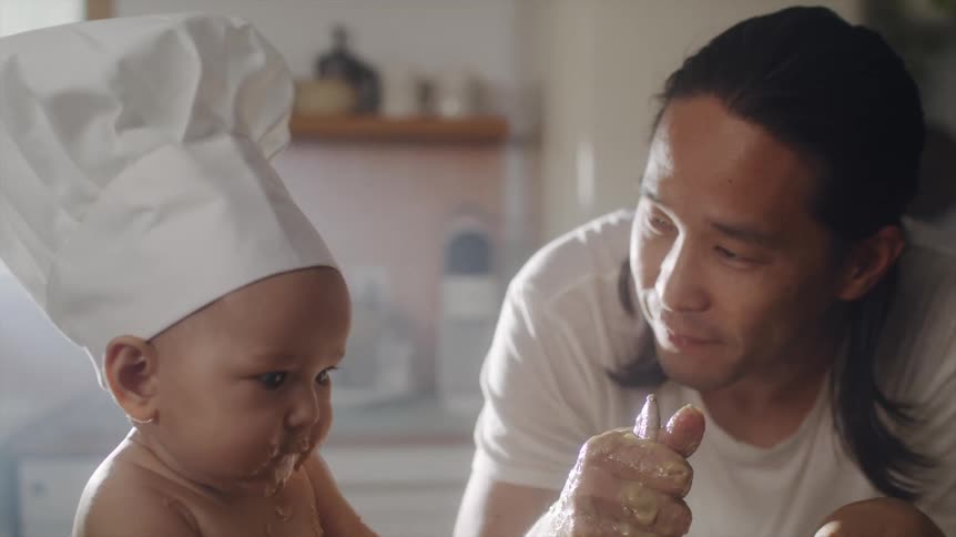 Asian man cooking playing with and kissing his baby son who is sitting on the kitchen counter wearing a chefs hat