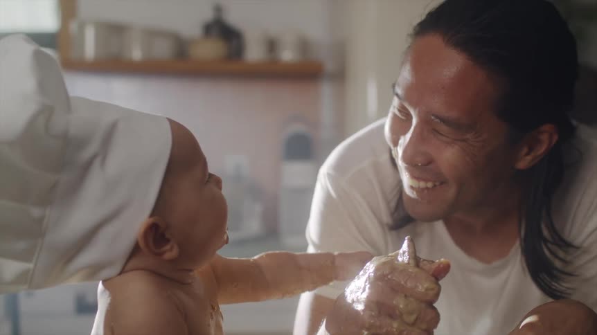 A man mixes batter as a baby touches his face in a home kitchen during the day