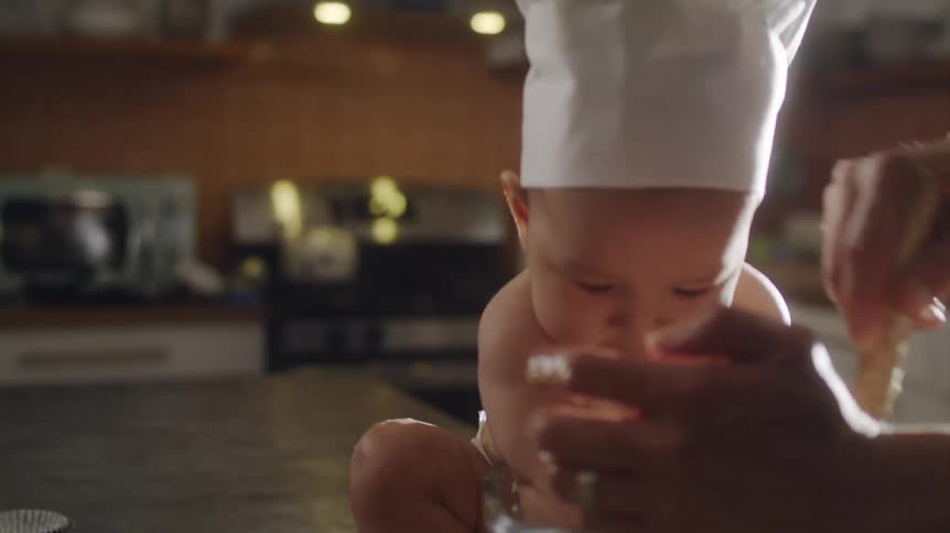 Baby son with chef hat and diaper plays with bowl while father stirring to make muffin on kitchen island in the kitchen