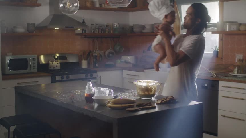 A man cooking in a kitchen picks his baby up from the counter