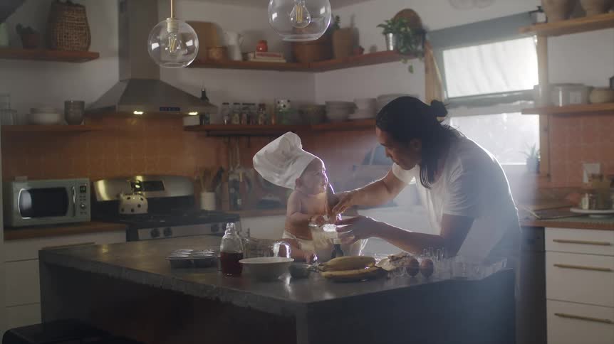Father stirs while baby wears chef hat sits on kitchen island and holds mixing bowl