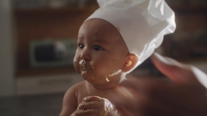 A baby wears a chefs hat and is covered with food while eating with its father