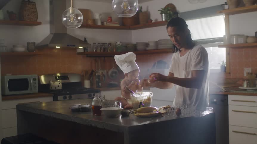 Father cracking an egg and mixing it in a bowl while his baby son is sitting and eating something next to him at the kitchen counter