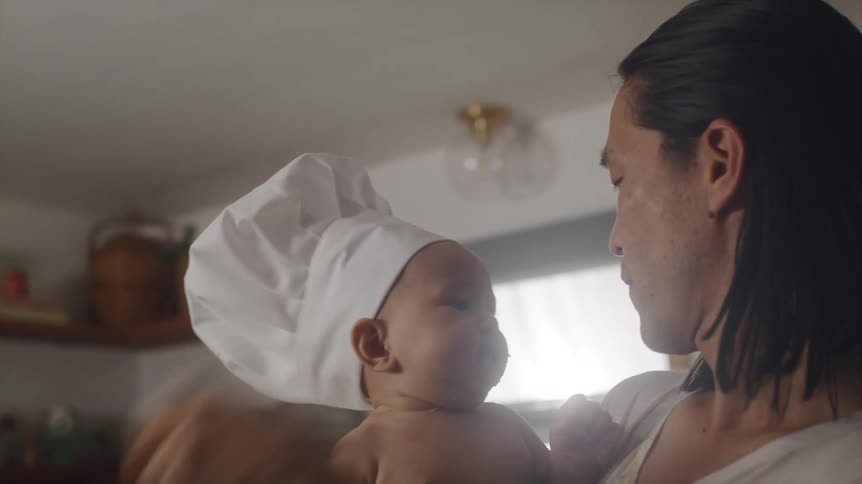Father holding his baby that is wearing a chef hat and cleaning his face with a hand cloth at the kitchen