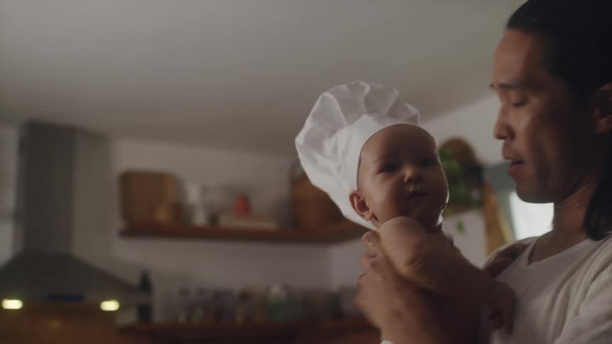 Father picking up his baby son wearing a chef hat and holding it in his arm at the kitchen
