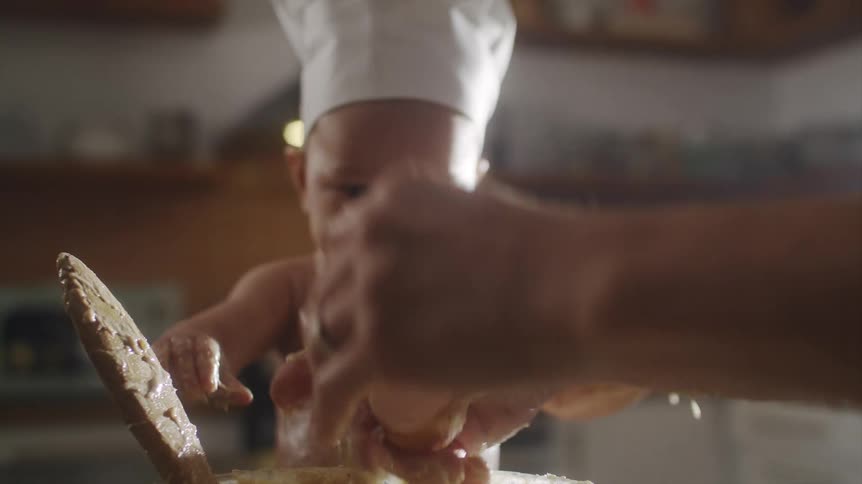 Man cracking an egg throwing it into a bowl and mixing it all while a baby wearing a chef hat is sitting right next to it