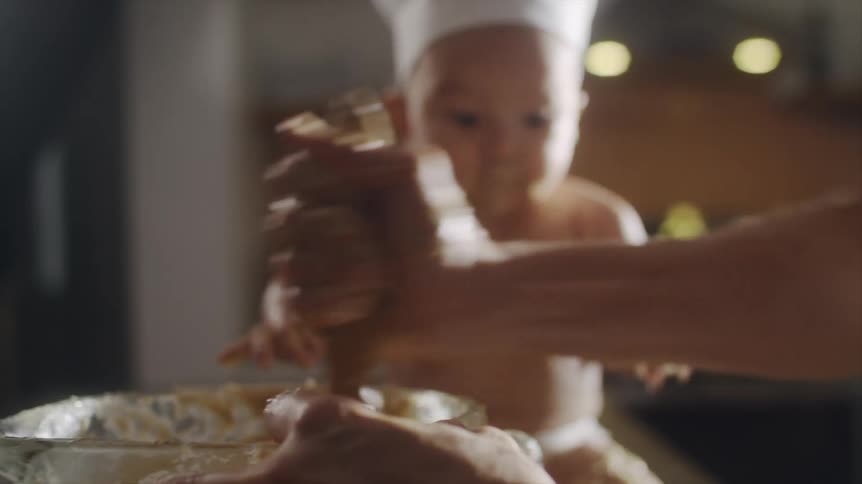 Batter is being stirred in a large bowl as a baby wearing a white hat sits nearby