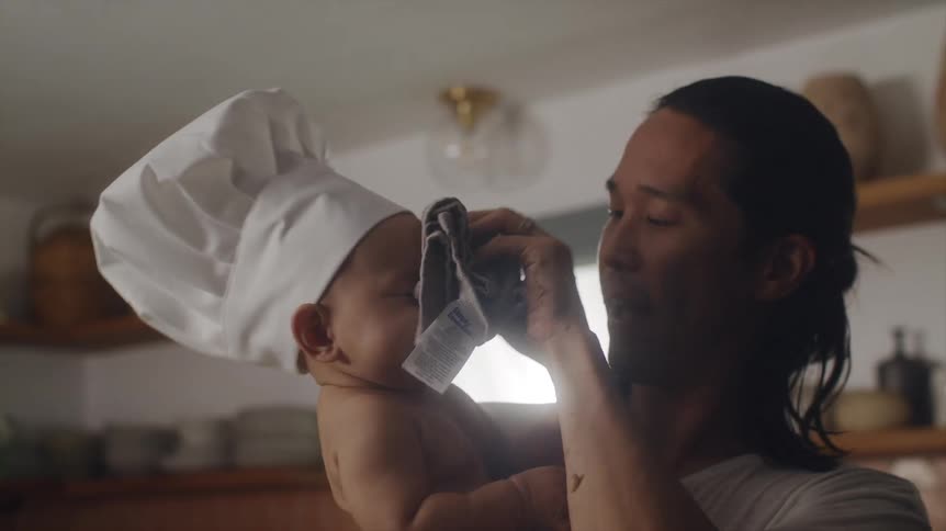 Father holding his baby son that is wearing a chef hat and cleaning his face with a hand cloth
