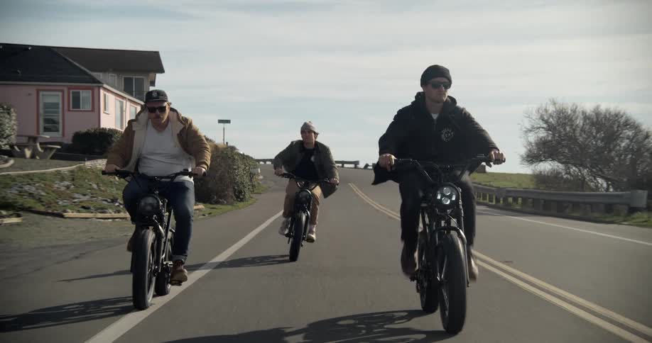 Three men are riding bikes on a street and pass by houses as an automobile travels in the distance behind them