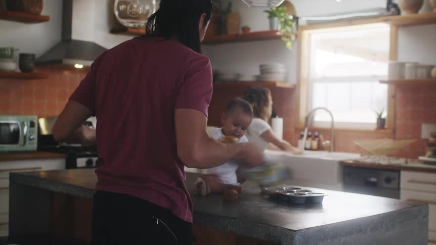 Father with long hair and red t shirt plays with baby son sitting on kitchen island with mother doing the dishes in the background