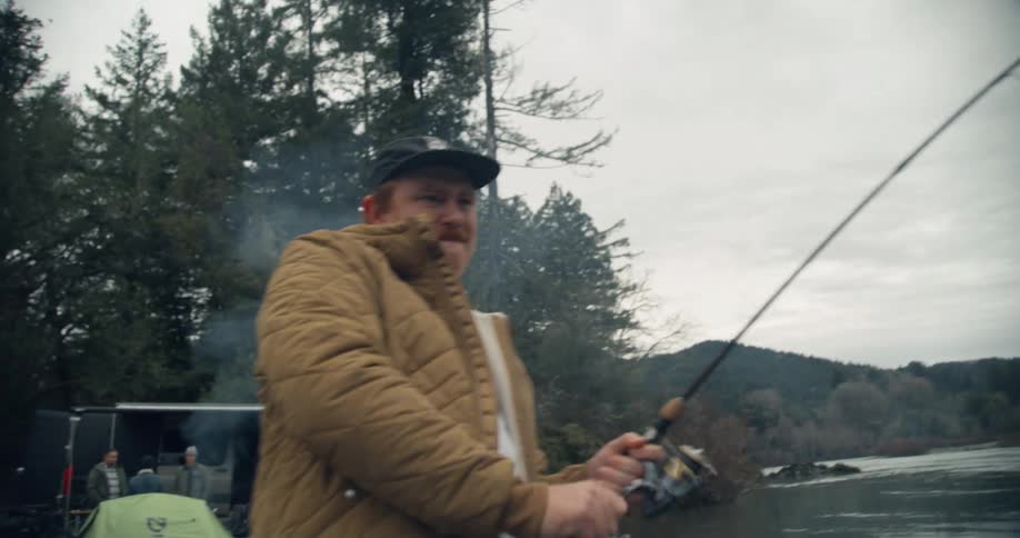 A man is fishing on the side of a lake and he gets a bite reels in the line
