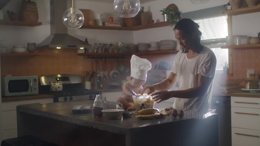 Man mixing food in a glass bowl at the kitchen counter while his baby son wearing a chef hat sitting next to it