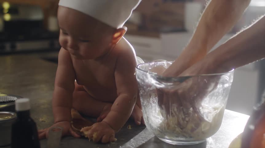 An adult mixes dough in a mixing bowl while a baby eats dough from a mixing spoon on the counter nearby