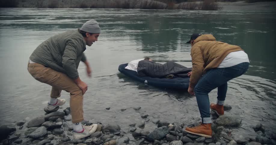 A man is lying on an air mattress and two men are carrying him to the lake and he is floating on the water